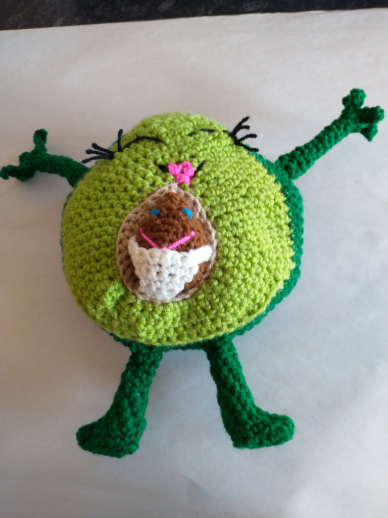 A crochet avocado with arms flung open and sporting big smile and pouted lips. The avocado seed in her middle is a baby in a nappy