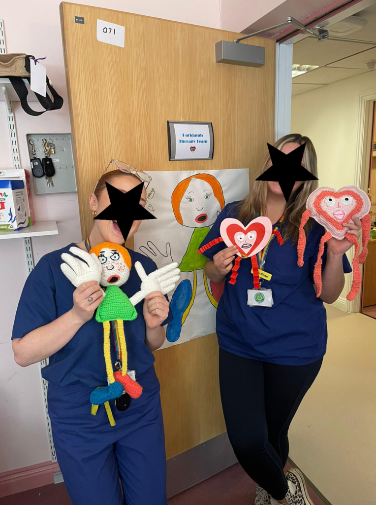 A photo of two health workers with their faces obscured by black stars. One is holding a toy of a lady with big white hands and large blue and orange feet and orange hair. The drawing of the lady is on the door behind her. The other is holding a cutout drawing of a pink heart man in her right hand and the crochet toy of the same in her left.