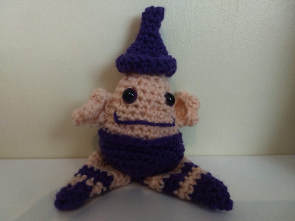 A photo of the crochet "wizard" in cream and purple. His head is cream, had has a very short body with a purple pair of shorts, two large pointed purple striped feed with purple tips, two large cream ears. no arms, and a purple pointed hat. He is smiling broadly