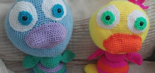 A photo of two crochet ducks sitting next to one another on a cream chair. The duck on the left is blue and green with green wings, blue face, blue feet and a purple beak. He has large blue eyes on white and a green set of three feathers on his head. The one on the right has a yellow head, orange beak, pink head and wing feathers, green eyes and yellow feet