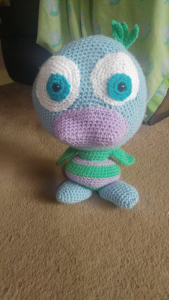 A photo of the blue, green and purple crochet duck standing on a beige carpet