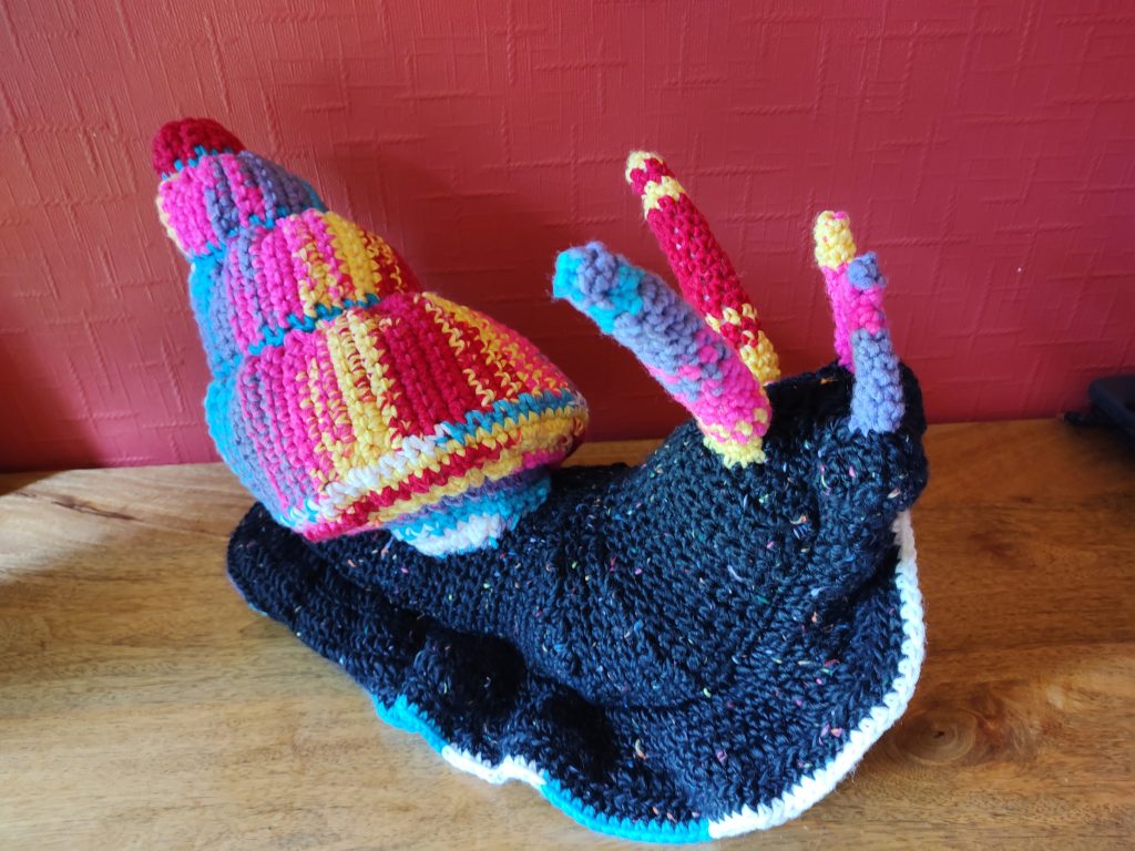 A crochet giant snail with a black, colour-flecked body and multi-colour shell and antennae in red, purple, yellow and pink.