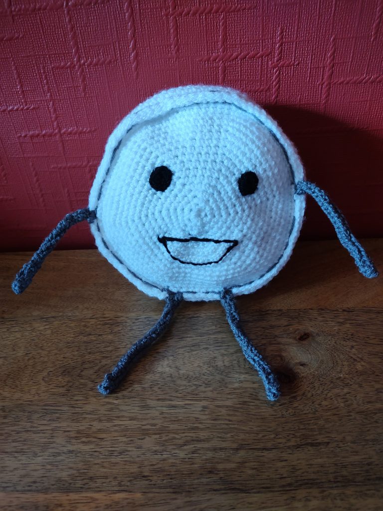 A white round crochet face with two round black eyes and a happy smile outlined in black. The face has two very thing grey arms and legs