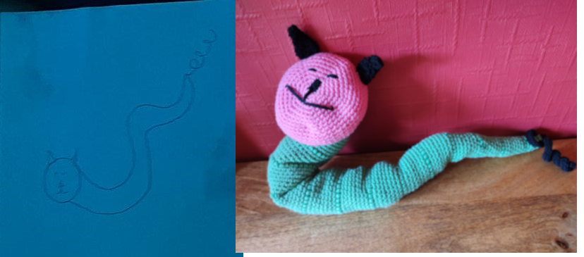 A pencil drawing of a worm or caterpillar on blue paper next to the crochet toy of the same on the right. The toy has a pink round face with little eyes and a big smile and black ears. The body is green and there is a black curly bit on the end of the tail