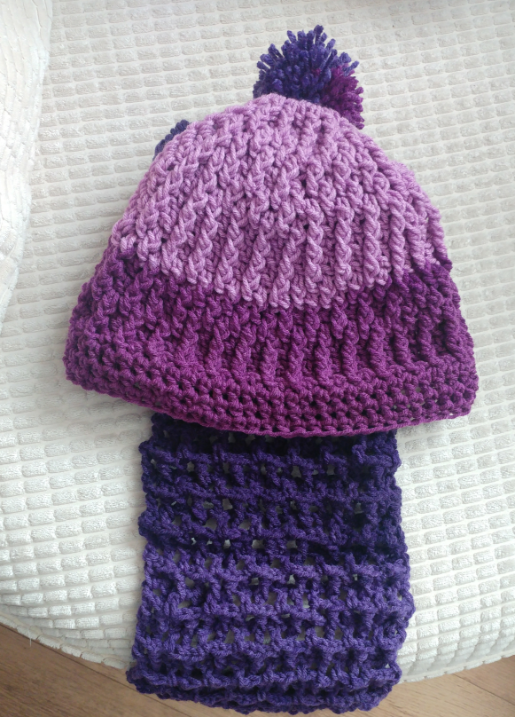 A photo of a variegated purple ribbed crochet hat and scarf. The hat has a pom pom on the top