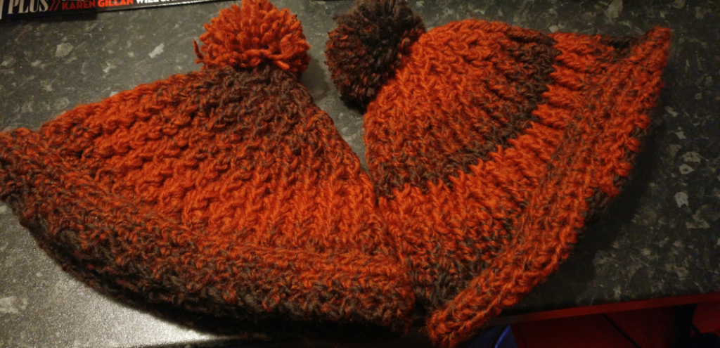 A photo of two red and very dark brown variegated colour ribbed crochet hats with pom poms on the top