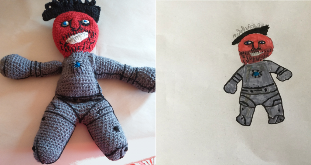 A photo showing a crochet pirate-like doll next to the child's drawing of the same, which actually may be supposed to be Iron Man. He is wearing a grey iron suit with a blue light in the centre. He has a very red face, a white grinning smile and a stubbly black beard. He is wearing a pirate like hat with grey hair on top. The crochet doll does not have the grey hair as the picture was taken before it was added.