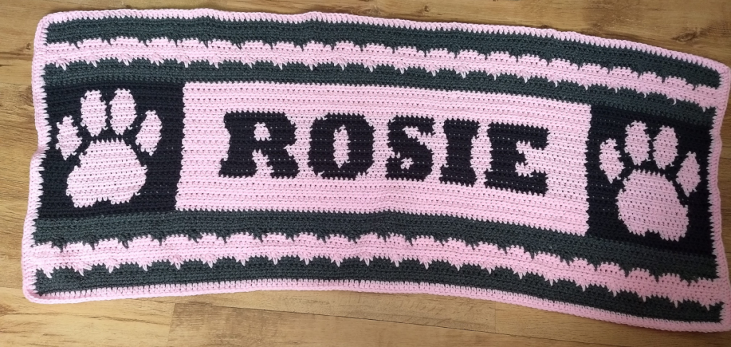 A crochet dog blanket in black and pink. The blanket has a pink central square with the name Rosie in it and two pink paws on black next to each side of the lettering
