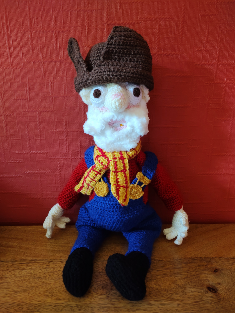 A crochet version of Stinky Pete from toy story. He has a brown miners hat on, a red long-sleeved top and royal blue dungarees with a red and yellow checked necktie. He has large eyes and nose and a fluffy white beard and moustache