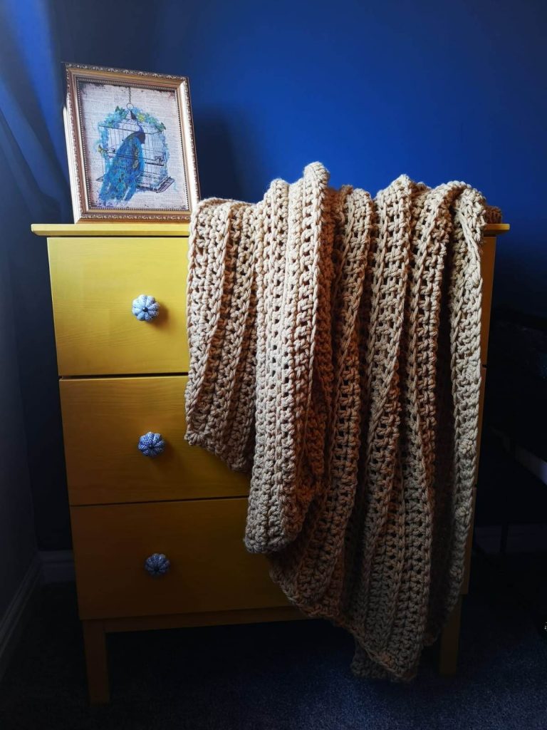 A tan crochet throw with a ribbed appearance which is shown in a blue room thrown over a wooden set of drawers.