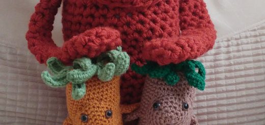 A photo of three crochet toys called Tree Thangs, which are basically long tree like toys with floppy hands and feet, leafy hair and round faces with two brown eyes and no nose or mouth. In this photo is a very large copper coloured Tree Thang with its hands on tow smaller ones about a third of its size which are caramel and light brown colours