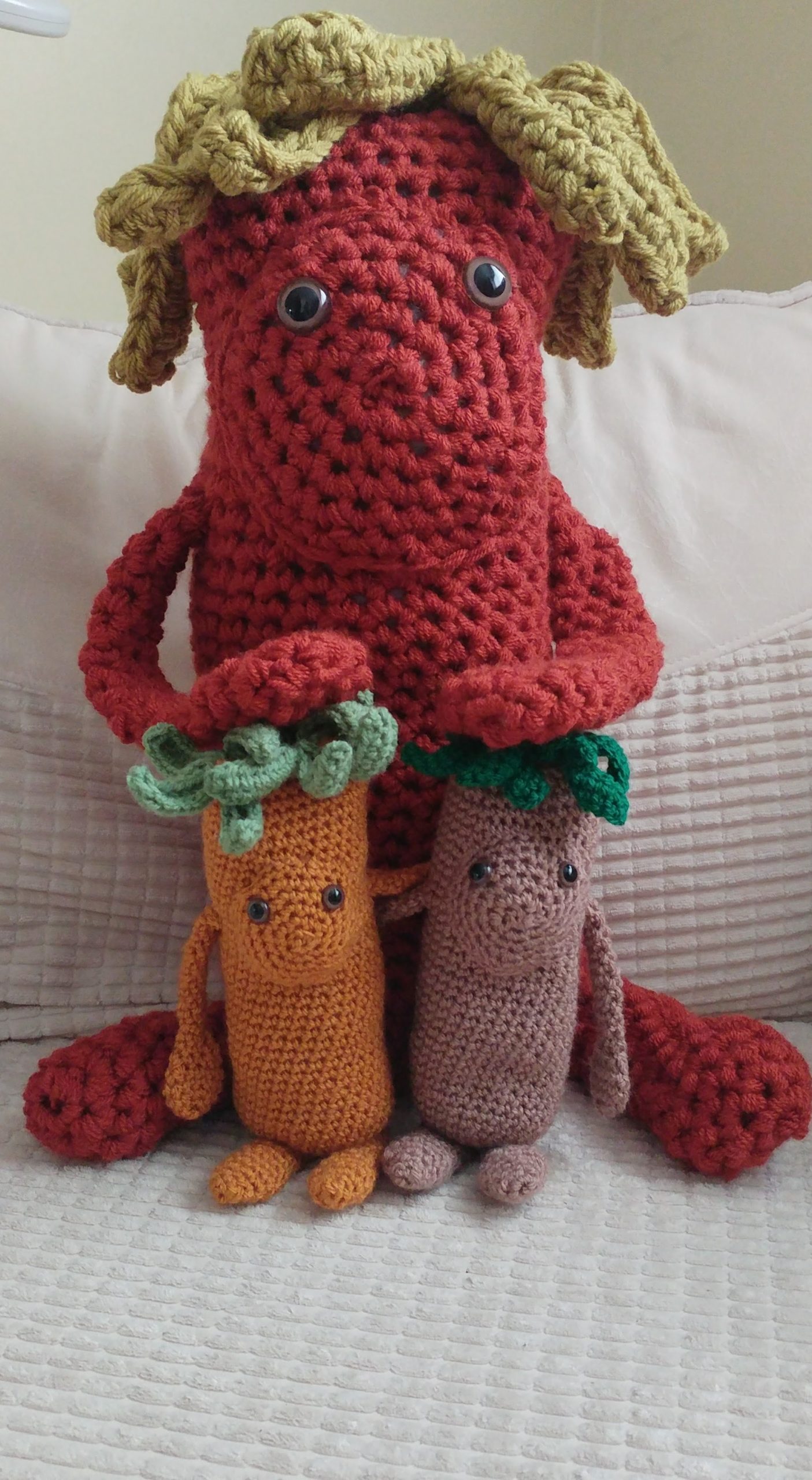 A photo of three crochet toys called Tree Thangs, which are basically long tree like toys with floppy hands and feet, leafy hair and round faces with two brown eyes and no nose or mouth. In this photo is a very large copper coloured Tree Thang with its hands on tow smaller ones about a third of its size which are caramel and light brown colours