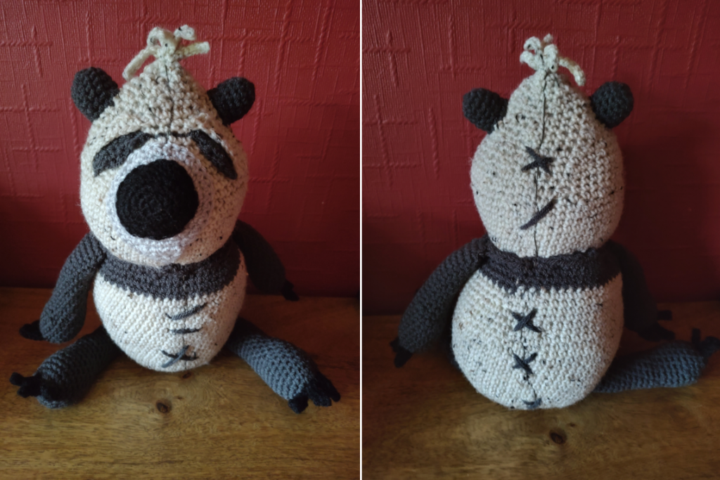 A photo of a bear made from tweedy yarn to give him a worn look. He has a big black round nose, dark brown eyes and ears, legs and arms and a dark brown strip under his head, which is cream coloured as is the rest of his body. He has crude sewing down his front and back in dark brown yarn. He has a fringe of tendrils on the top of his head