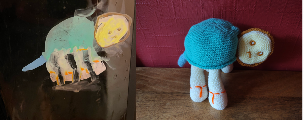A painting of a turtle with a yellow head, yellow legs, blue tail and green shell on a window. The crochet toy is on the right.