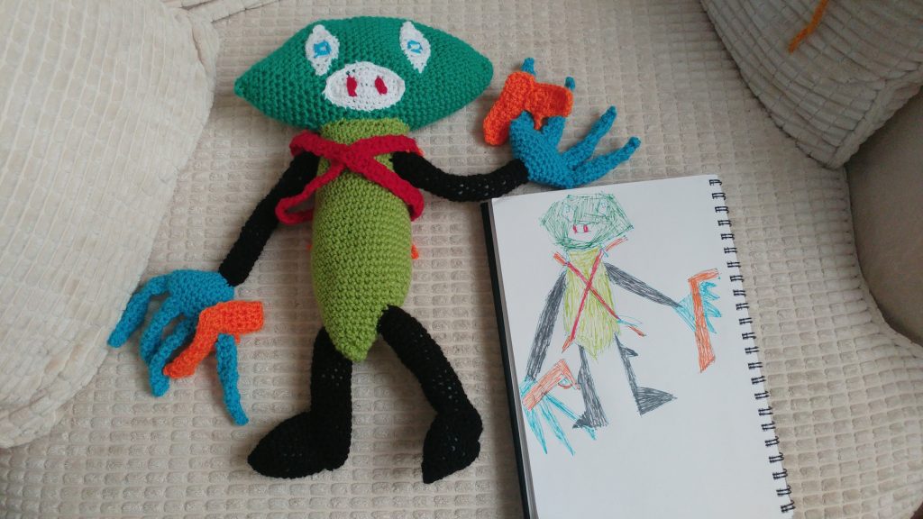 A crochet monster next to the drawing pad on which the child has sketched it. The monster has a diamond shaped green head, white diamond eyes with blue circles and a white oval mouth with two red teeth. It has a green body and is wearing red criss-cross bands. It has black legs and arms and blue hands with four long fingers. It has an orange gun in each hand