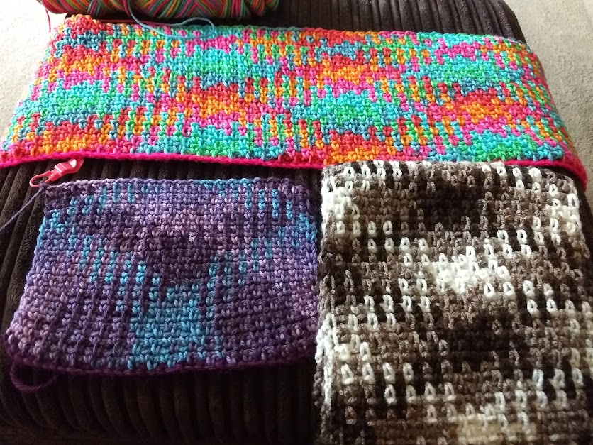 A photo showing three crochet works in progress with argyle patterns. On the top is a colourful piece in blue, green, pink, yellow and orange. Underneath to the left is a piece in shades of purple and blue and on the right one in shades of brown and cream