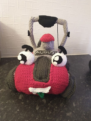 A crochet lawnmower toy. the front is red with a dark grey stripe in the middle and he has two large white eyes on top and a wide smile with a green tongue sticking out. The wheels are dark grey and he has a light grey handle with a black grip and power cord coming off it.
