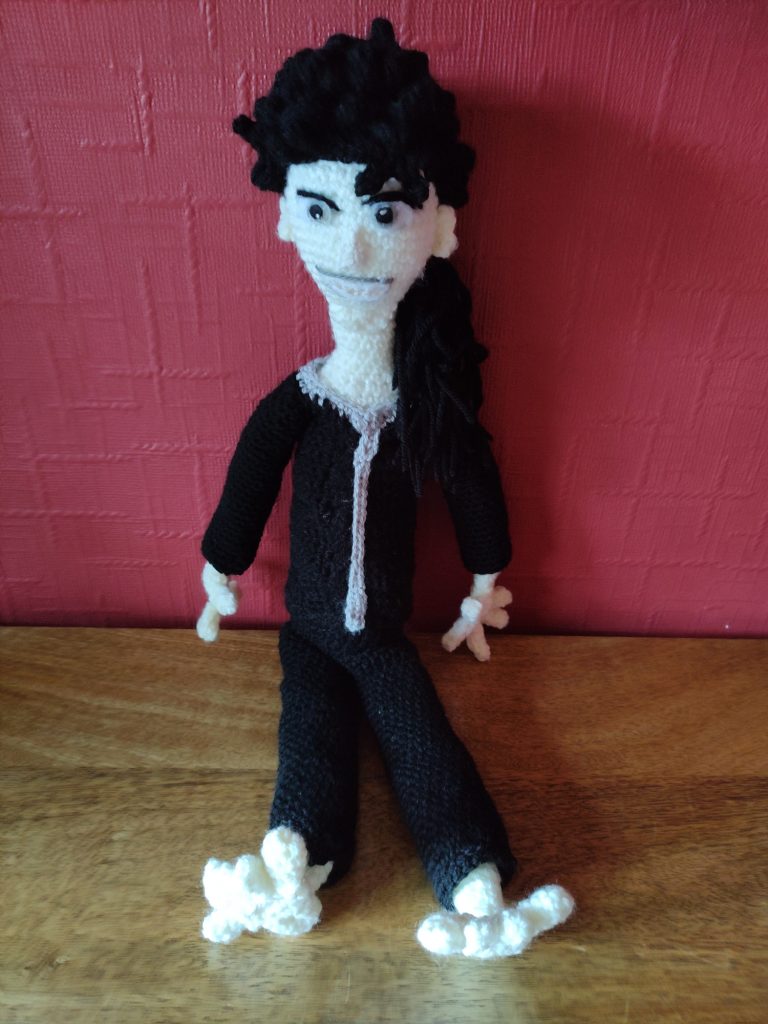 A crochet doll of a manga character. He has pale skin and bare feet and is wearing a black tracksuit with a grey stripe down the front of the top. He has long, curly black hair, very dark eyes and grey lips