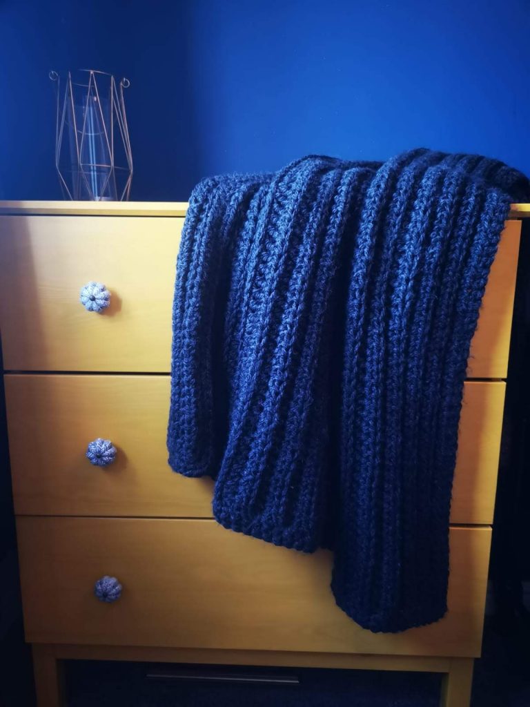 A blue crochet throw with a ribbed appearance which is shown in a blue room thrown over a wooden set of drawers.