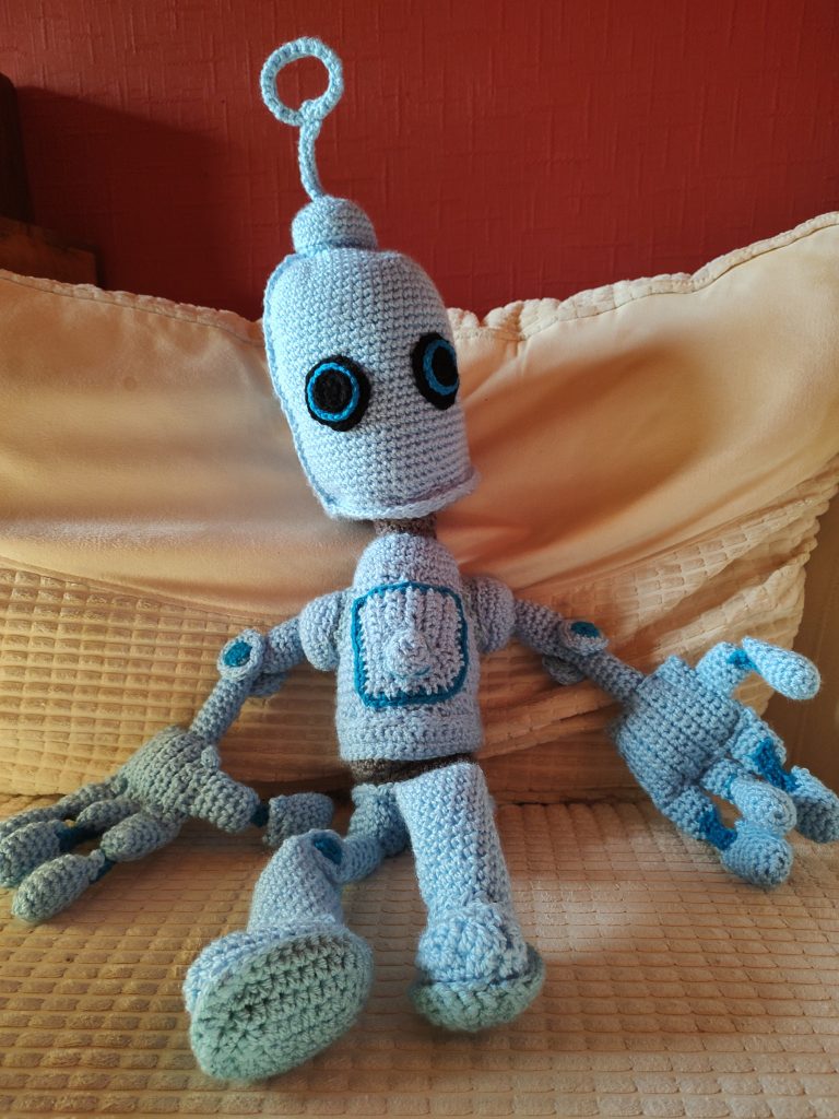 A photo of a crochet blue robot sitting on a cream chair. He is a light blue with darker blue around his chest place and between his finger joints. He has a large round black eyes rimed with the darker blue and has a light blue ring antenna on the top of his head.