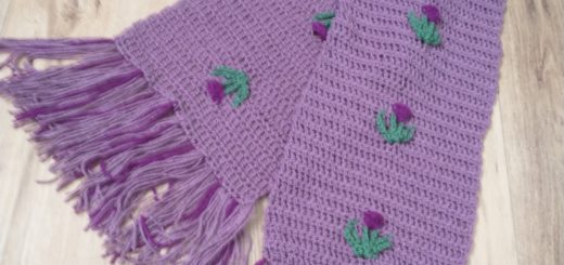 A photo of a purple crochet scarf with thistles in darker purple and green evenly spaced up the centre. The scarf has a fringe made up of the lighter and darker purples yarns