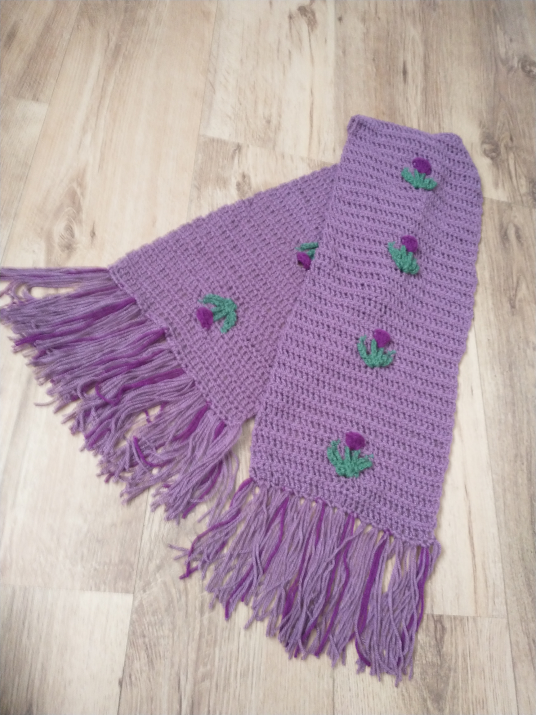 A photo of a purple crochet scarf with thistles in darker purple and green evenly spaced up the centre. The scarf has a fringe made up of the lighter and darker purples yarns