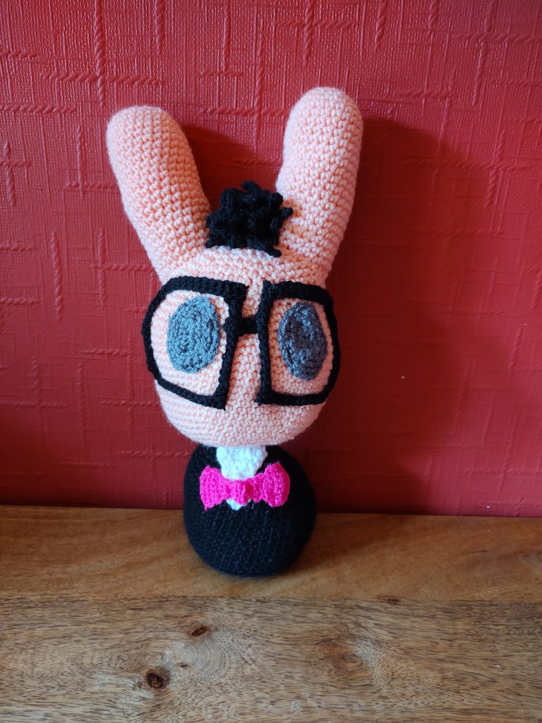 A photo of a crochet bunny based on a game character. he has a very large peach coloured head and long ears, a much smaller body wearing a black tuxedo and a bright pink bow tie and very large square glasses with thick black rims. He has solid oval dark grey eyes and a tuft of black hair between his ears. He doesn't have arms or legs