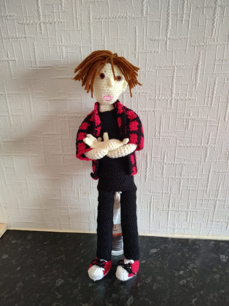 A crochet doll of a manga character. This one has short brown hair and is wearing a red and black checked shirt over a black t-shirt and black, tight, trousers. He has brown eyes with dark pupils and is standing with his arms folded. He has red and white sneakers on.