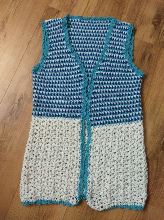 A crochet blue and white cotton sleeveless waistcoat. The top part is blue and white mixed and the bottom is white. There is a light blue edging around the waistcoat