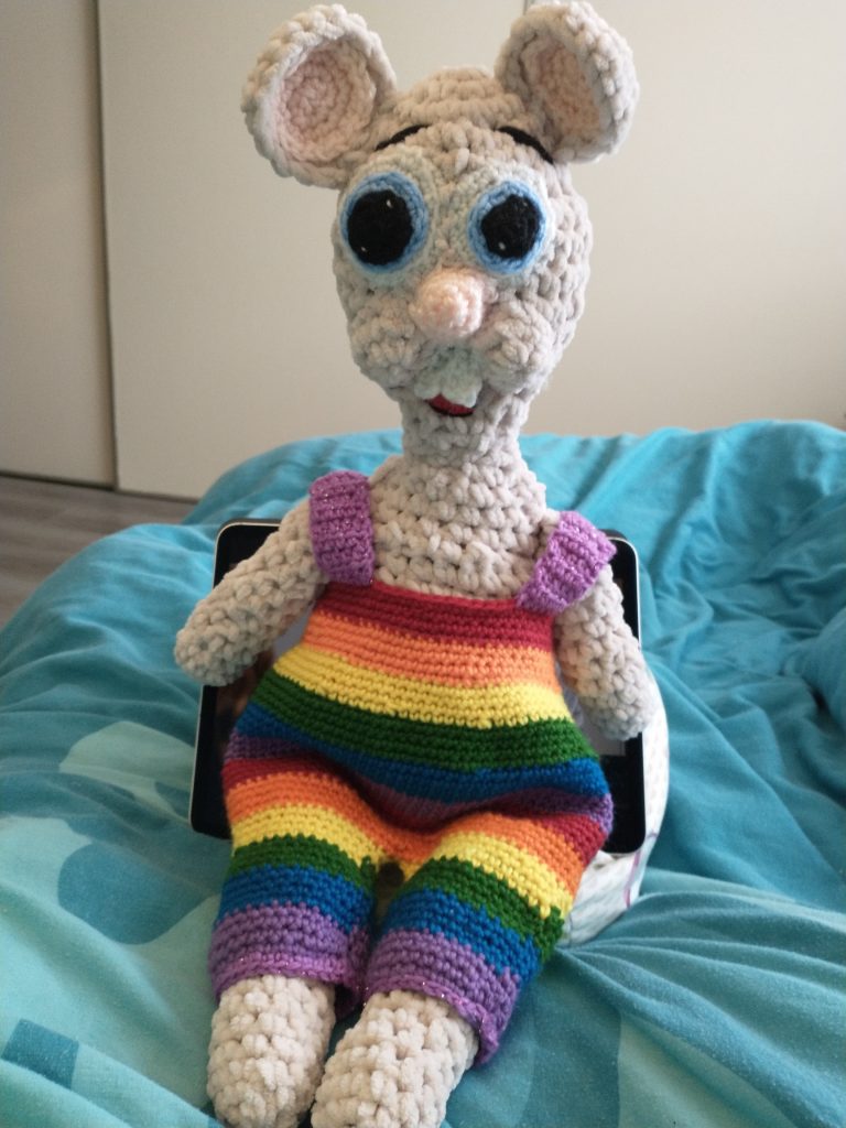 A tan crochet mouse with crazy looking blue eyes with huge black pupils wearing a pair of dungarees in rainbow colours