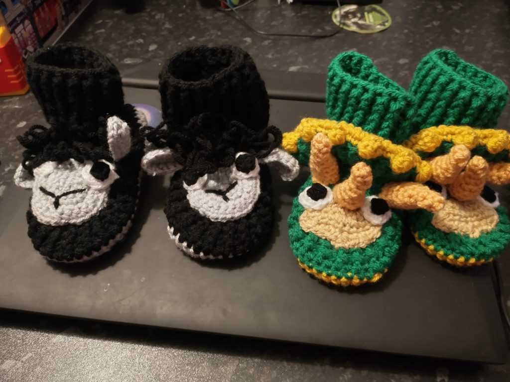 Crochet animal slippers. On the left is a pair of black sheep slippers with white faces. On the left is a pair of triceratops slippers in green with orange-yellow horns and faces and yellow neck frills