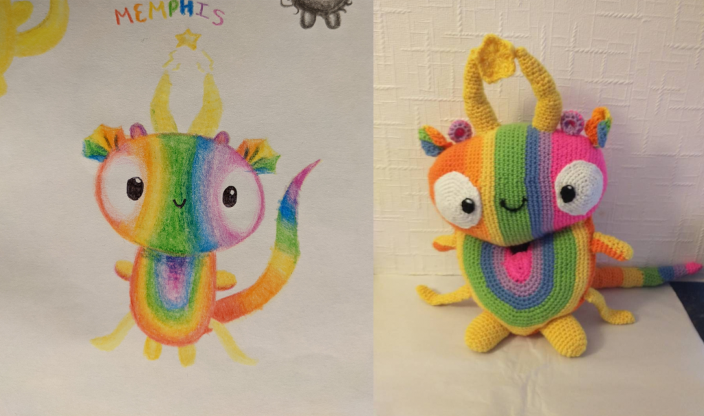A photo of a drawing of a "Dewd", which is a rainbow creature with a rainbow oval body with the colours going out from pink, through lical, blue, green and yellow. The oval head goes from pink on the right to orange on the left. It has two large yellow horns with a star between them, two large round white eyes with highlighted black pupils and a small black smiley mouth. The crochet animal is on the right. It also has two yellow and orange arms, two orange and yellow tentacles at the bottom and two yellow legs and a rainbow tail.
