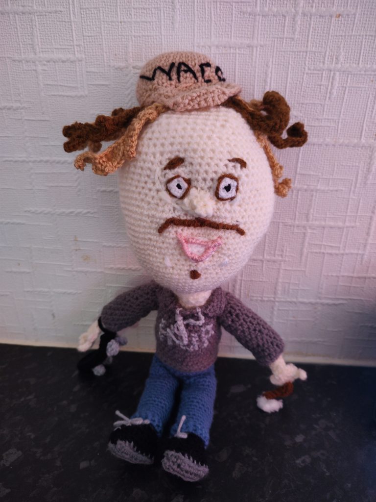 A photo of a crochet Egg Man, with a very large egg-shaped cream head in relation to his body. He has on a tan baseball cap with the letters WACO on it and his "hair" is a mixture of dark yellow and brown yolk. He has small round white eyes rimmed in brown, brown eyebrows and a thin brown moustache. He is smiling. He is wearing a grey sweatshirt blue jeans and black trainers