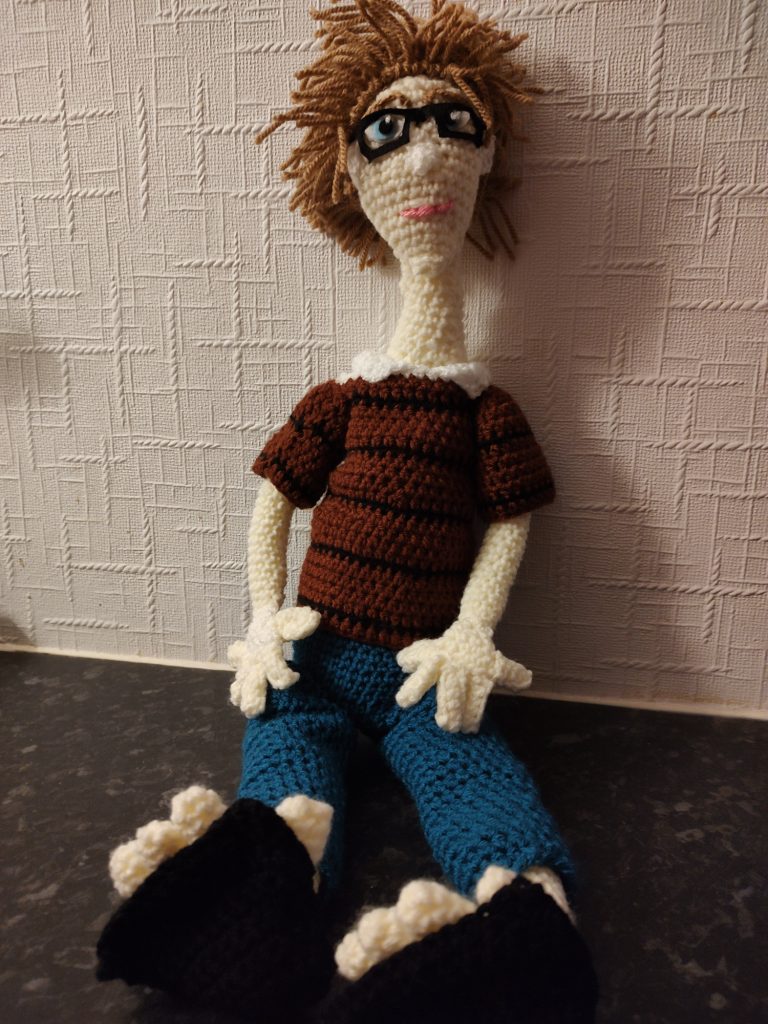 A crochet doll of a manga character. this one is wearing blue trousers and black triangular flip-flops. He has on a brown jumper with thin black stripes and is wearing a white shirt under it with the collar sticking out. He has wild brown hair sticking up and blue eyes. He is wearing thick black square glasses