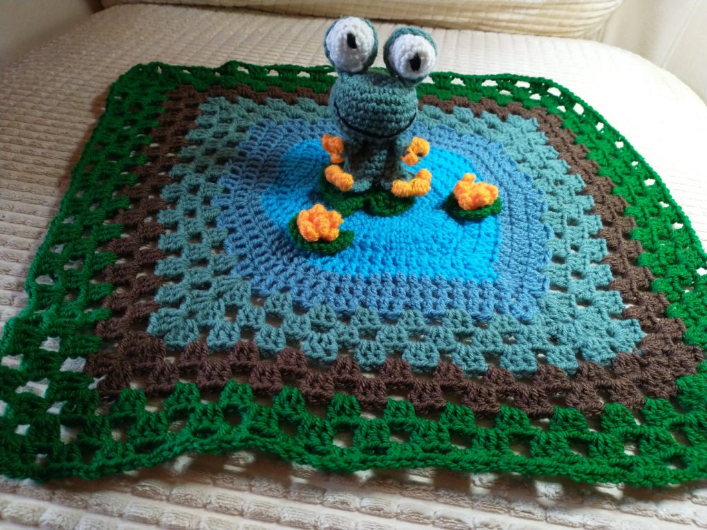 A green crochet frog sitting in the middle of a multicoloured granny square blanket meant to represent a lily pond. At the centre is a bright blue hexagon. Radiating out from that is a darker blue hexagon and then squares in blue, brown and green. The frog is sitting on a green lily pad and is surrounded by three lily pads sporting yellow-orange flowers.