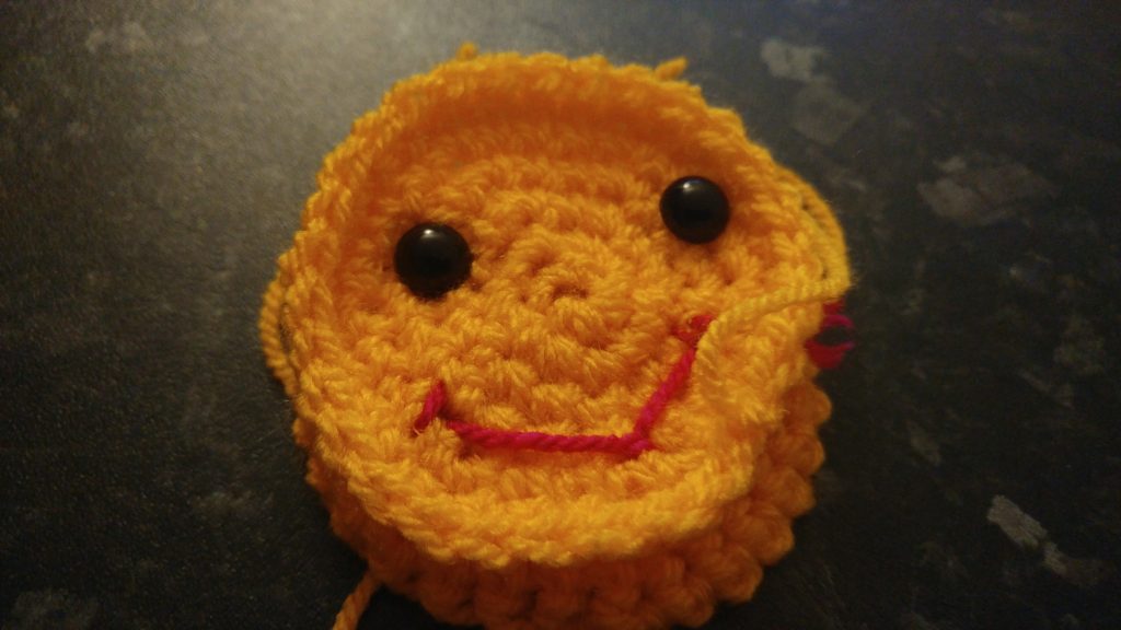 A photo of a yellow crochet happy face with two black toy eyes and a smiley little red mouth. The face is still being worked on