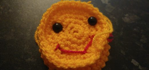 A photo of a yellow crochet happy face with two black toy eyes and a smiley little red mouth. The face is still being worked on