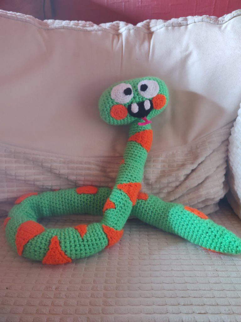 A crochet snake from the kid's program Hey Dugee. The snake is sitting curled on a white chair and is bright green with orange markings. It has two orange cheeks, large round white eyes and a smiling black mouth with two white teeth. A little forked pink tongue is poking out from the mouth