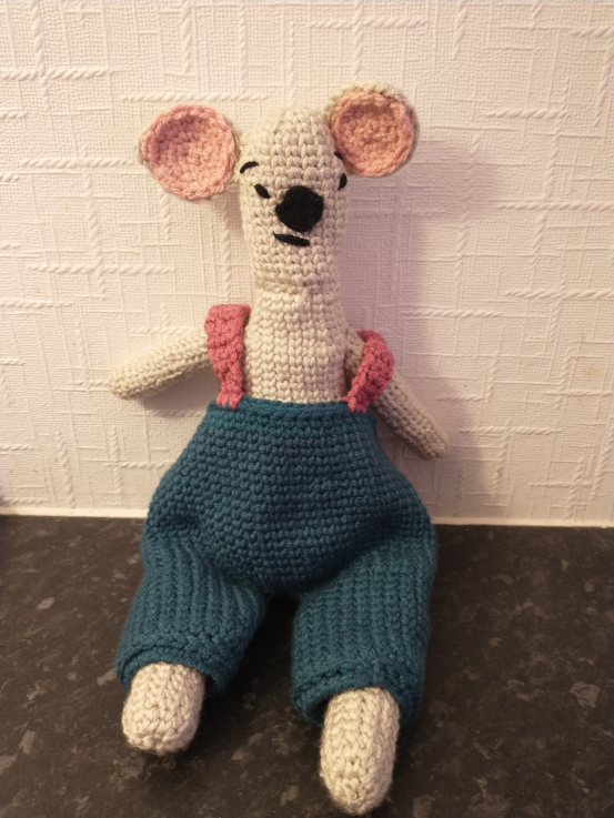 A large crochet toy standing on the floor next to a white kitchen cupboard which he is almost the height of. He is made with tan, fluffy yarn, has very large black eyes, long floppy earns and very chunky arms and legs