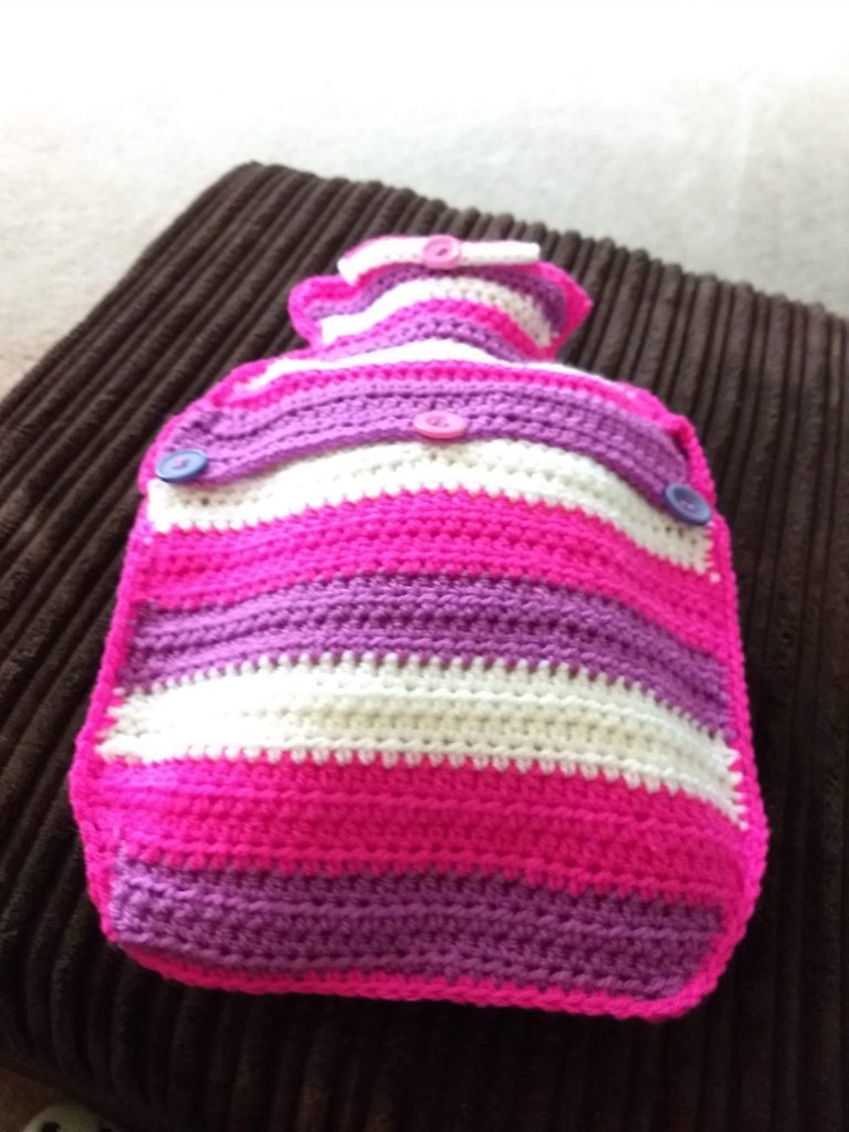 A hot water bottle with a crochet cover in bright purple, pink and white stripes