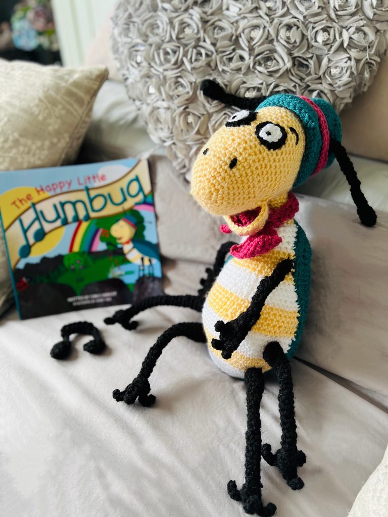 A photo of a crochet humbug next to the book that inspired it. The humbug is yellow with a white and yellow underside, green wings and black legs and antennae. It is wearing a green bowler hat with a pink ribbon around it and is wearing a pink bow tie.