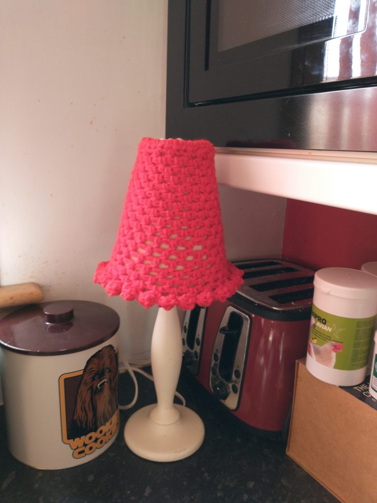 A photo of a red crochet lampshade with bobbles around the bottom sitting next to a red toaster, a black microwave and a Wookie Cookie biscuit tin