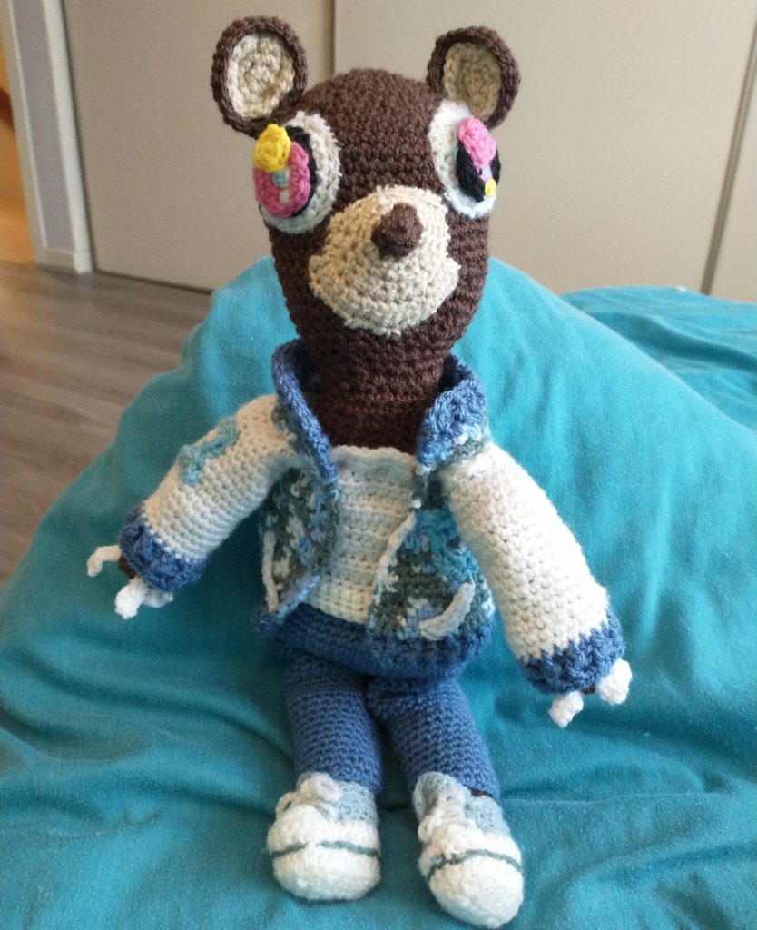 A photo of a dark brown crochet bear with eyes with pink, black, blue and white circles and a blue and grey camouflage jacket, blue trousers and white sneakers, sitting on a blue bedspread