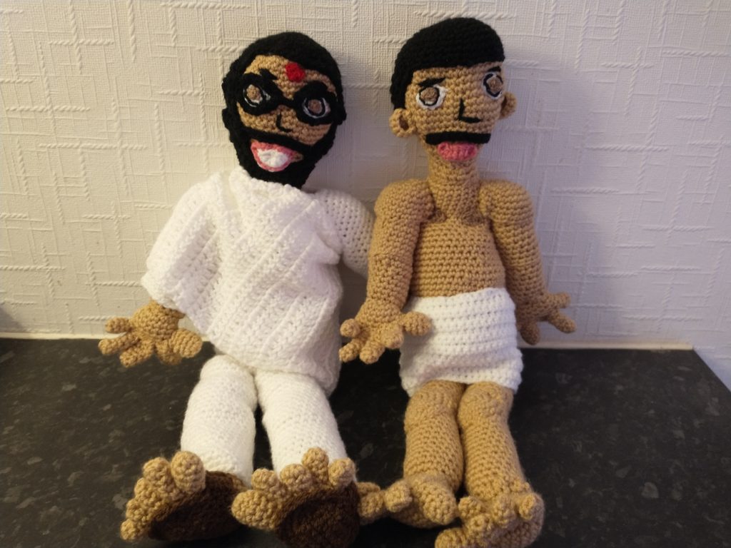 A photo of two crochet dolls representing gurus. The one on the right has a bare torso and legs and is wearing a wrap around his waist. He has black hair and a black moustache. The one on the left has black hair and a black moustache and beard and sports black glasses and a red dot on his forehead. He is wearing a wrap around shawl, white top and trousers and sandles