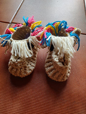 A pair of crochet llama slippers sitting on a terracotta tile floor. The slippers are beige with a cream fringe on the front and blue, pink and yellow coloured fringes around the sides