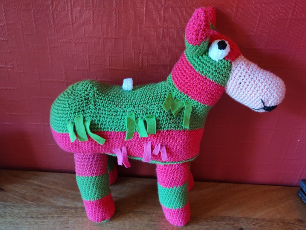 A pink and green striped crochet donkey with strips of green and pink felt to make him look like a pinata donkey