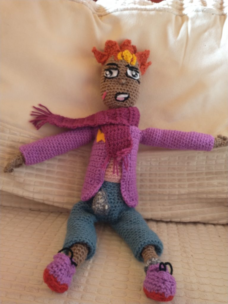 A crochet doll representing a manga character. He is wearing blue trousers and a pink top and purple jacket. he has on purple sneakers with orange soles and his hair is spiky and the same burn orange. He has on a purple scarf and has yellow markings on his forehead.
