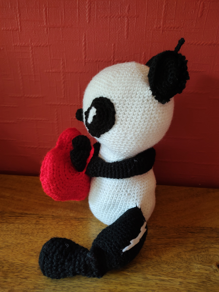 A side view of a crochet panda facing left. He has a white lightning flash visible down one thigh and is holding a red heart in his paws