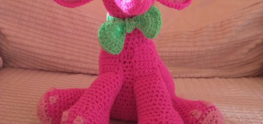 A photo of a pink crochet elephant sitting with his back legs splayed out and his front legs straight in front of him. He is bright pink with black eyes in white circles and has light pink foot pads, toes and inner ears. He is wearing a bright green bow tie