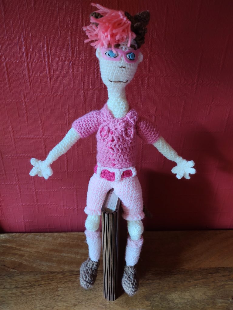 A crochet doll based on a manga character. He has a dark pink top with ruffles, light pink shorts, pink socks and brown boots. He has brown hair with a pink fringe in front and is wearing brown ears. He has pink goggles on and very blue eyes.